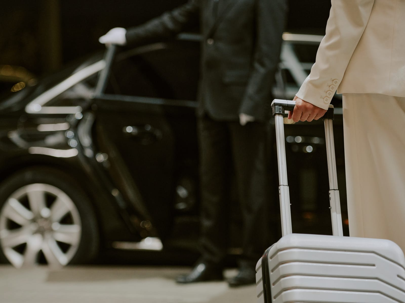 A driver holding open the door of a dark saloon car for a traveller with a suitcase.